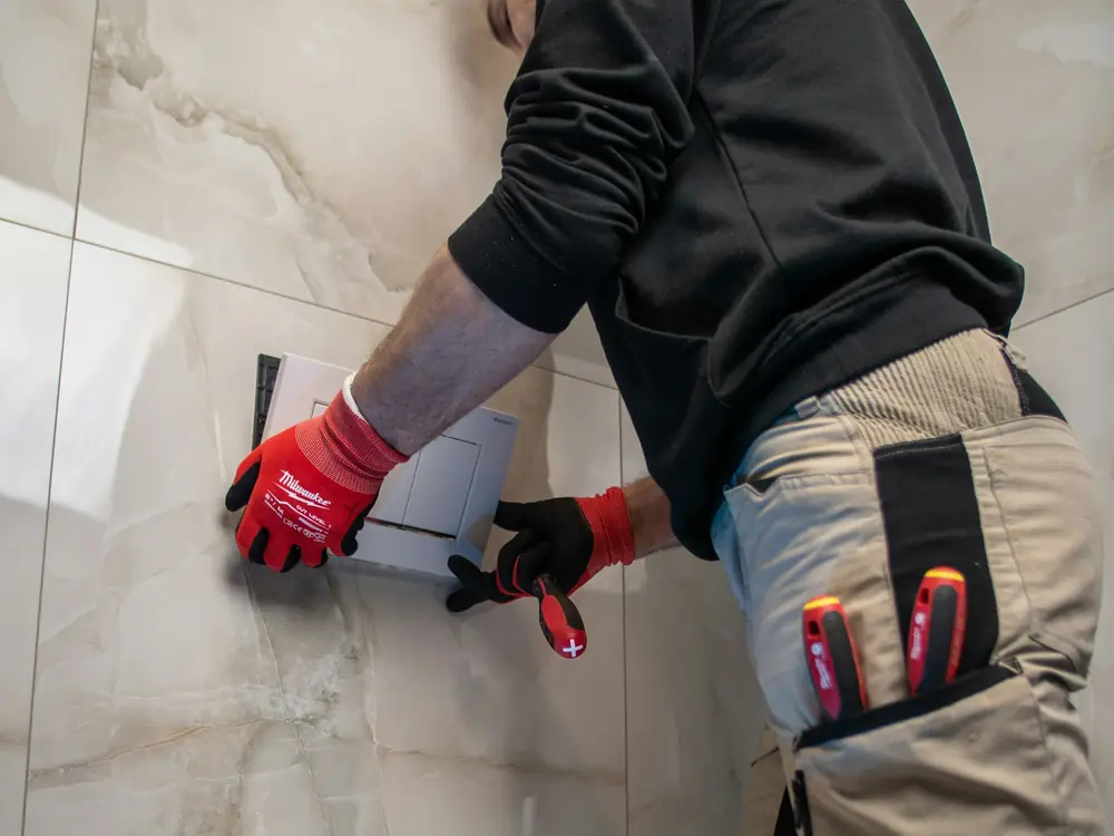A plumber in red gloves fitting a flush plate above a wall-hung toilet