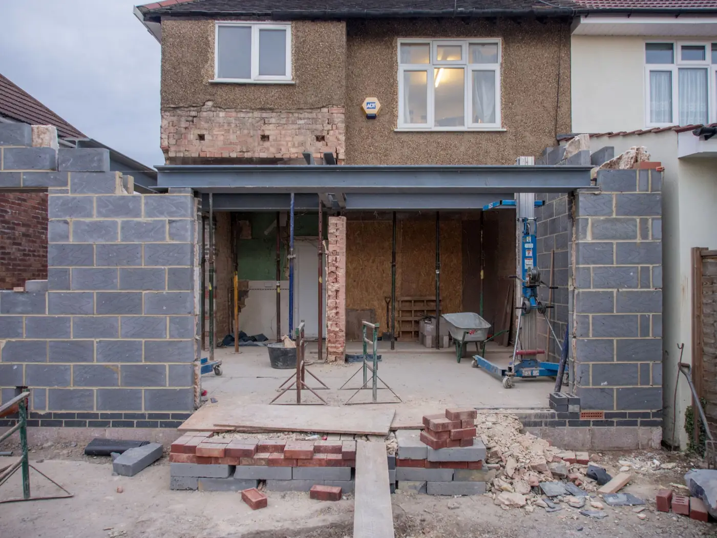 The back of a house opened up for an extension: new blockwork walls either side, a steel beam across the opening and bricks stacked on the slab