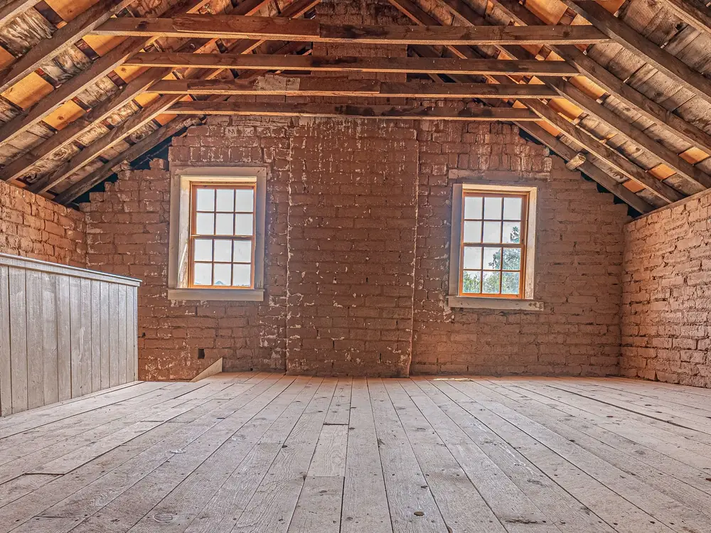 An empty loft with bare floorboards, exposed roof timbers and two windows in the brick end wall