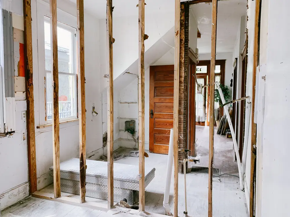 A house stripped back to its timber stud walls, with the staircase and a panelled door visible through the frame